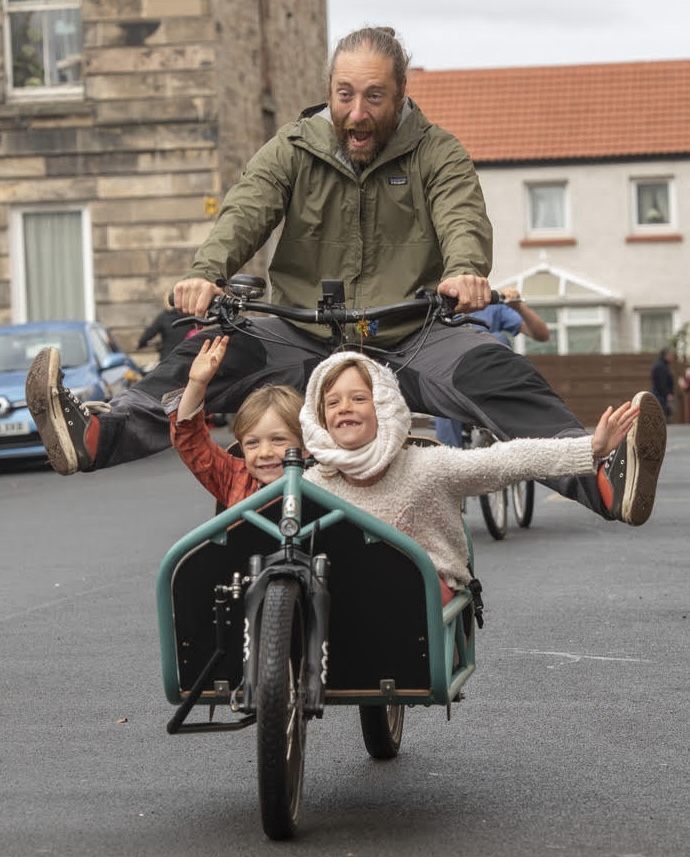 A man is riding a bike with two children in a basket at the front. They are all laughing. 