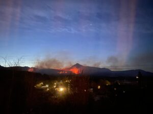 Photo of a mountainous landscape at dusk - a large area of a mountain in the distance is on fire with smoke rising from it